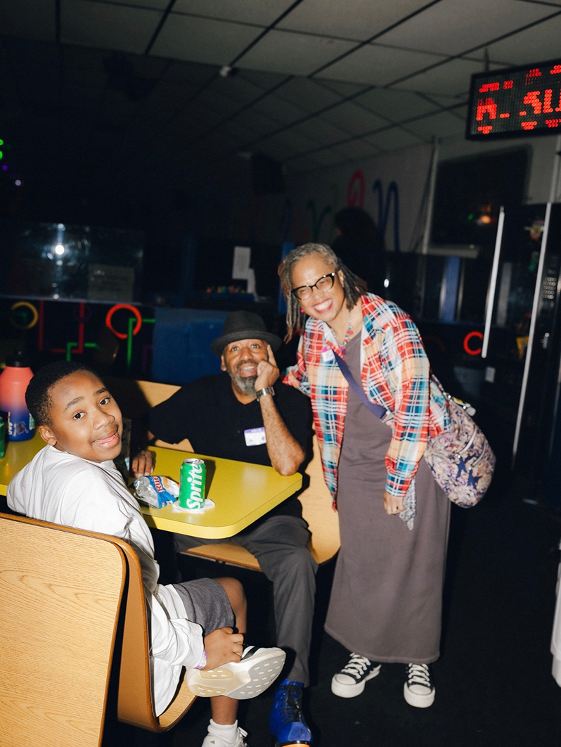 Three people smiling around a table in a dim arcade, with a neon-lit background and digital clock.