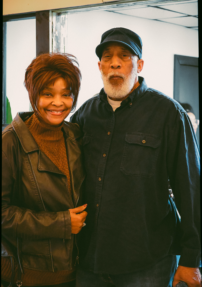 Two people standing indoors, smiling at the camera in dark jackets and hats.
