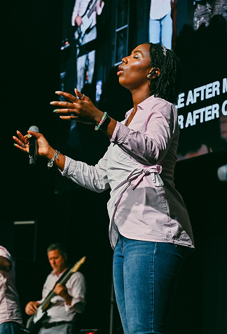 Singer on stage with eyes closed, hands raised, and guitarist in background against a dark screen