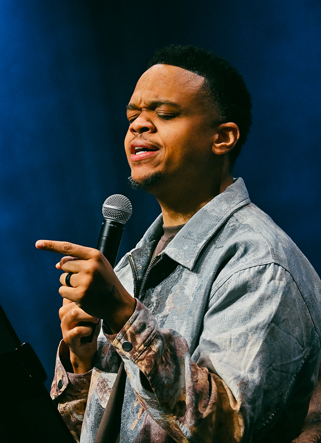 Singer holding a microphone and pointing, performing on stage under blue lighting