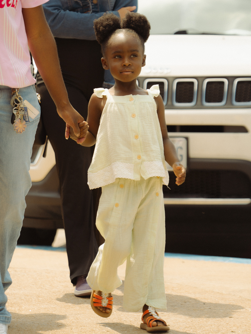 Child walking hand-in-hand with an adult beside a parked vehicle, wearing a white outfit and sandals.
