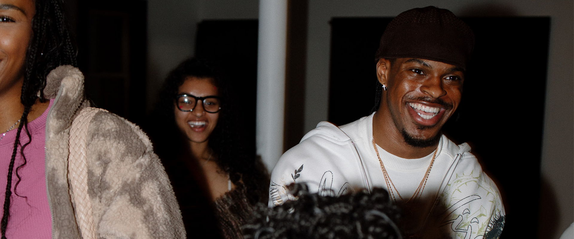 Smiling people at an indoor event, including one in a white shirt and brown beanie in the foreground