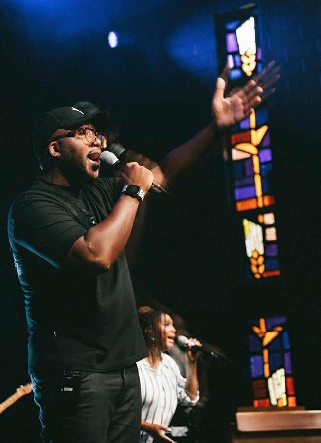 Singer performing on stage, microphone in hand, with colorful stained glass and blue lighting in the background