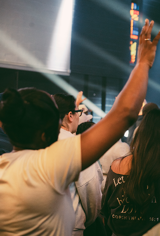 People with raised hands facing a bright stage or screen in a dim indoor crowd.
