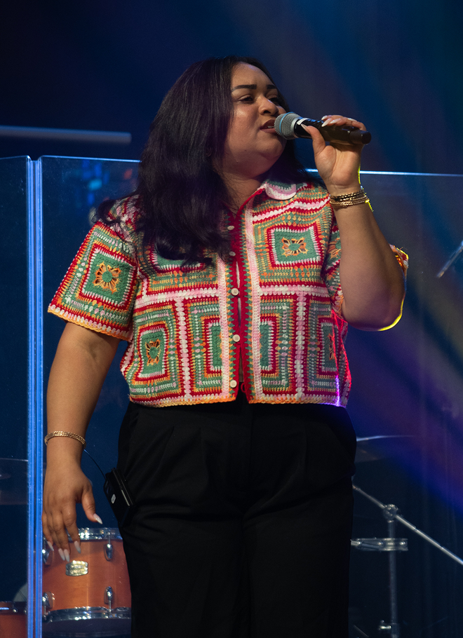 Singer performing on stage, wearing a colorful patterned top and black pants, singing into a microphone.