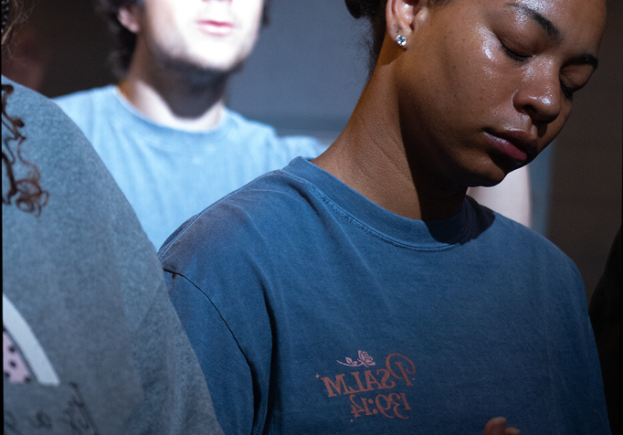 Three people in blue shirts sit in dim light, one in the foreground with eyes closed.