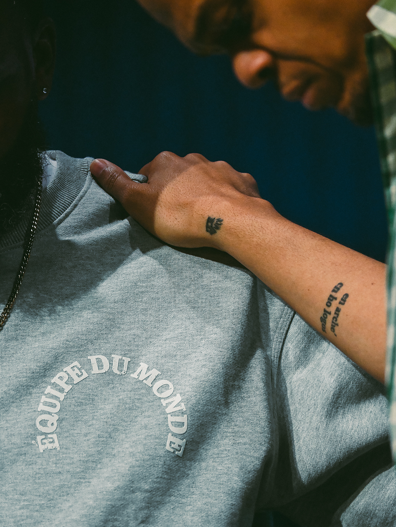 Person resting hand on a gray sweatshirt, with visible tattoos and a close-up of a face above.