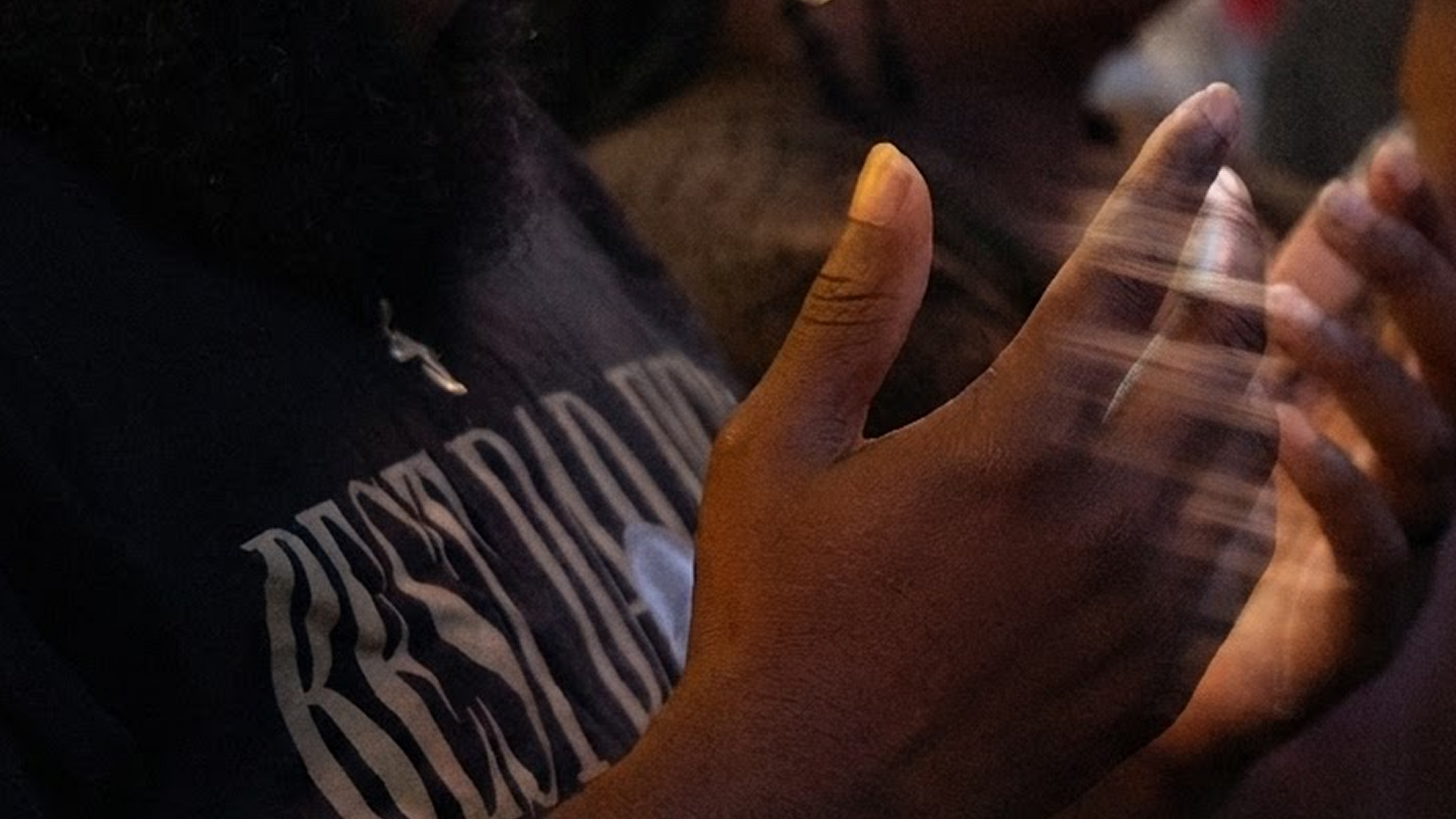 Hands clapping near a dark shirt with white lettering, lit by warm, low light.