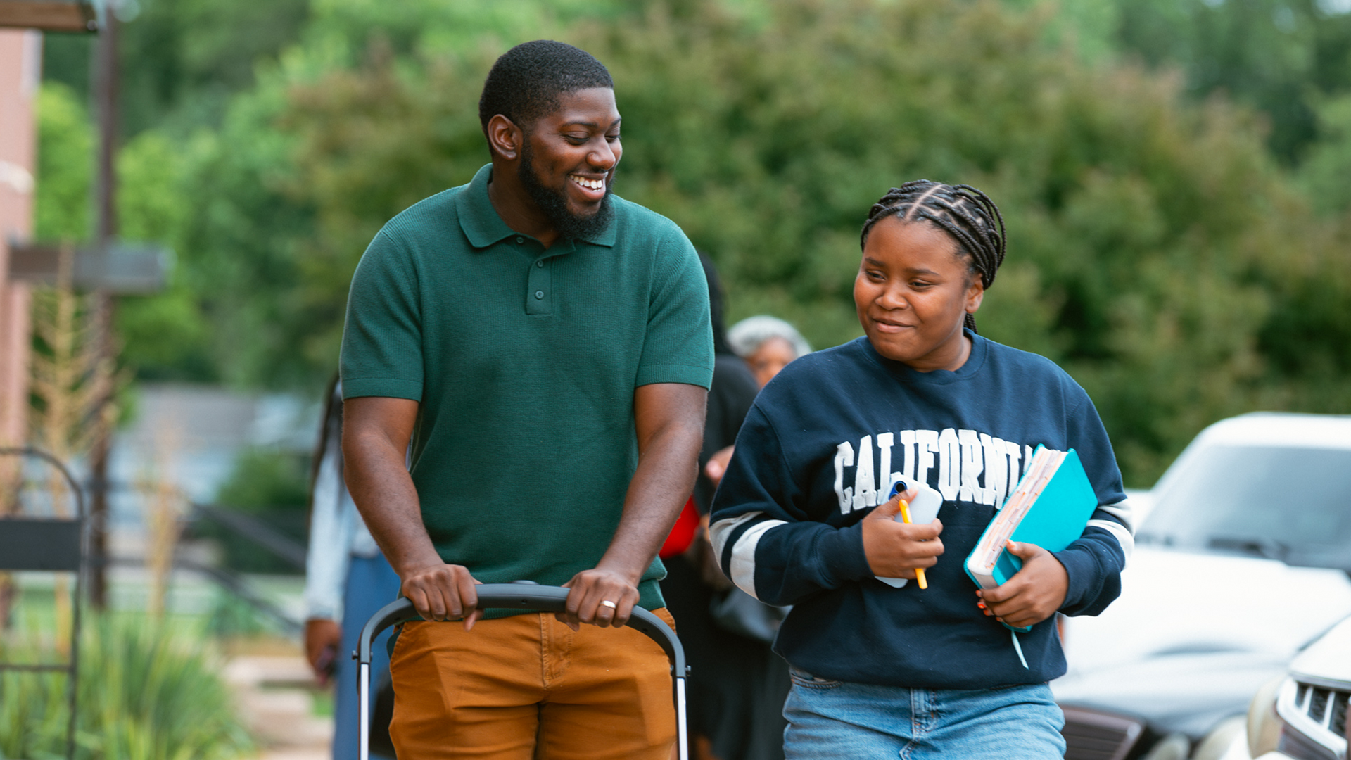 Two students walking outdoors, smiling and talking, one holding books and a notebook