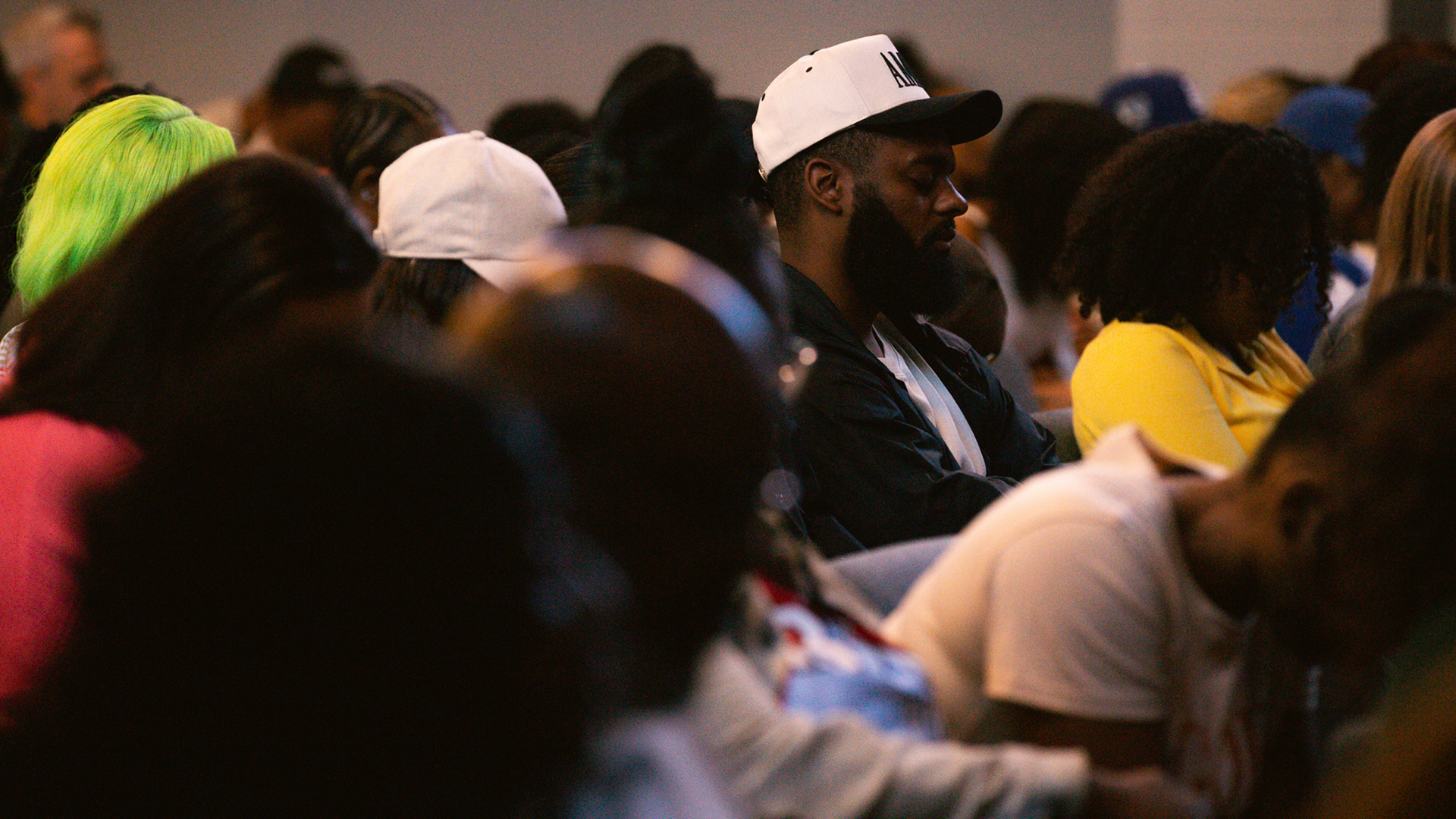 Crowd seated indoors, with a man in a white cap centered among blurred attendees