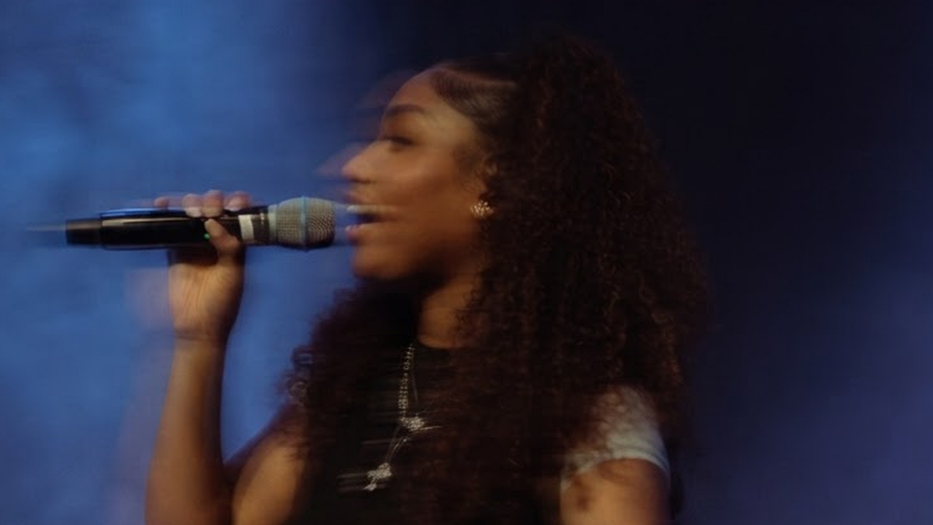 Singer performing on stage with a microphone, against a blue-lit background.