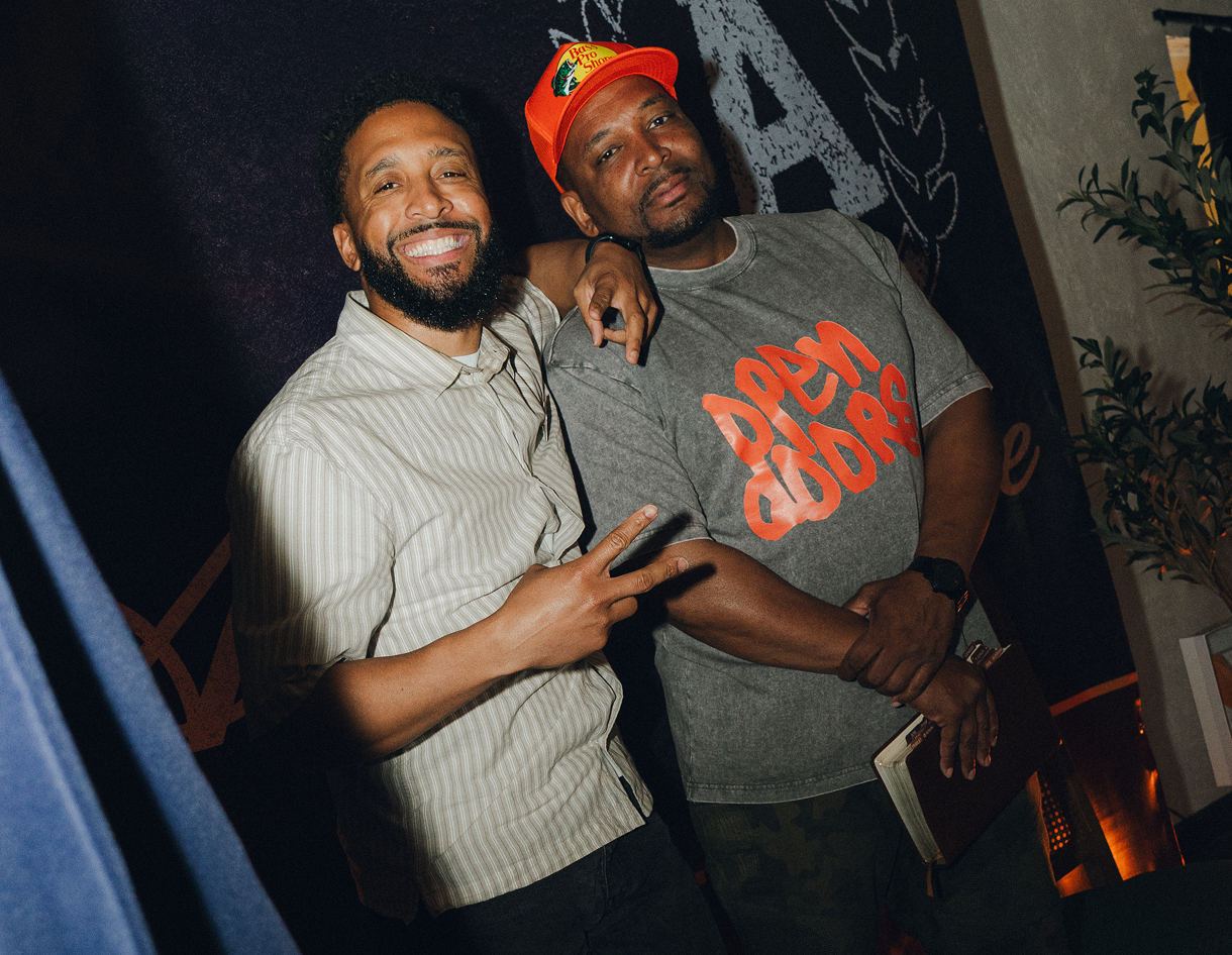 Two smiling people posing together at a dimly lit party, one holding a drink and making a peace sign.