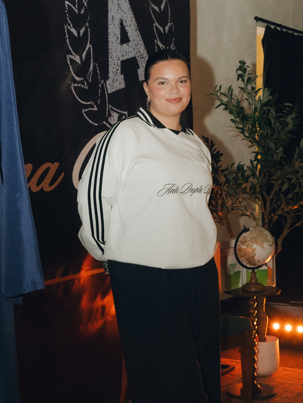 Person posing indoors in a white hoodie with black stripes, standing beside a lit plant and backdrop.