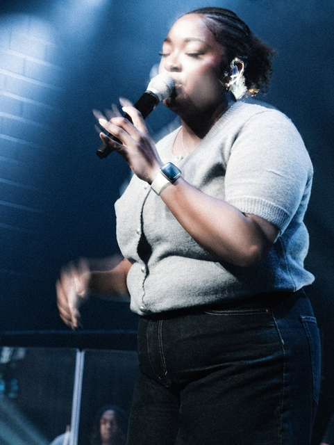 Singer performing on stage with a microphone, wearing a light gray top and black pants under blue lighting