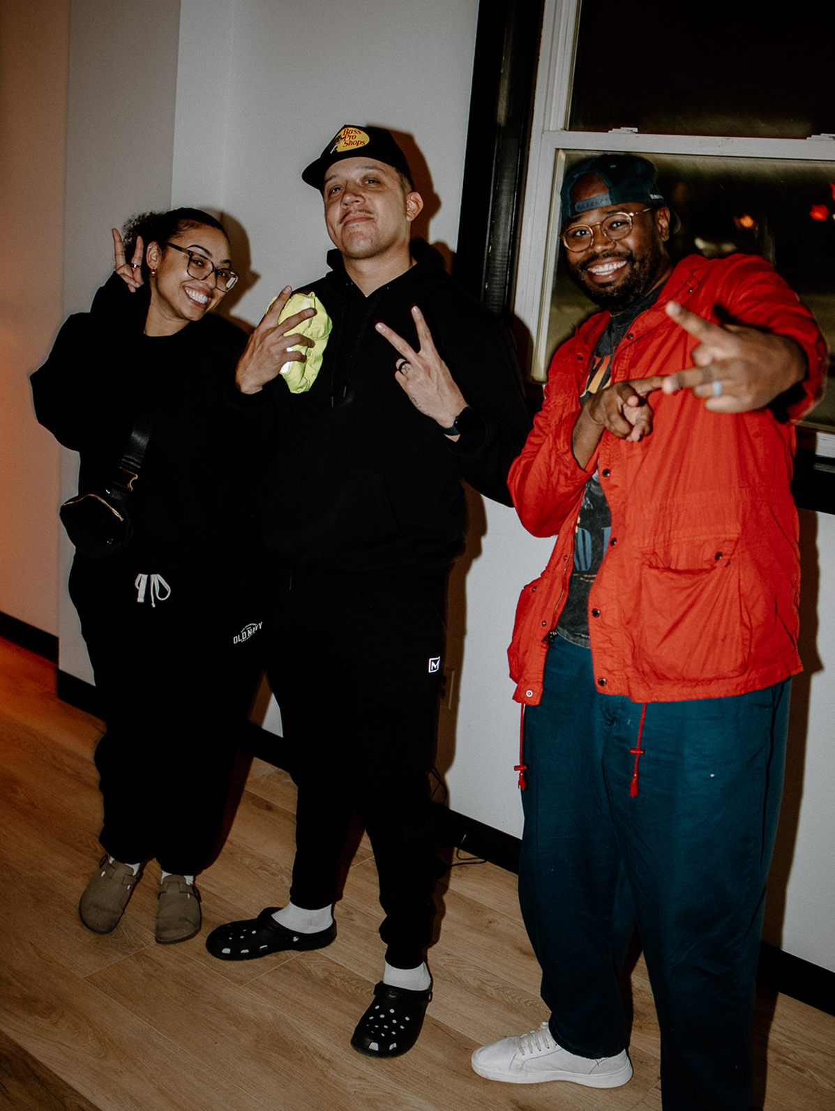 Three people posing indoors, smiling and making hand gestures, wearing dark and red outfits.