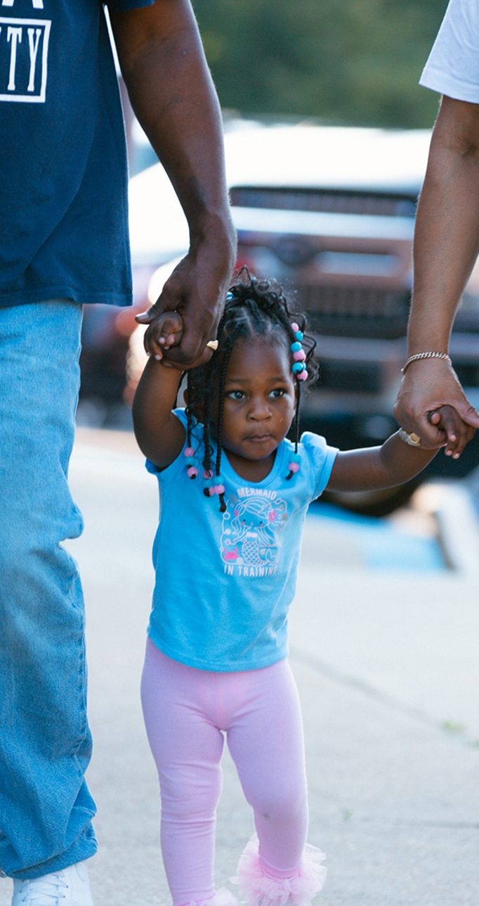 Two adults holding a toddler’s hands while walking outdoors, with plastic bags and colorful clothing.