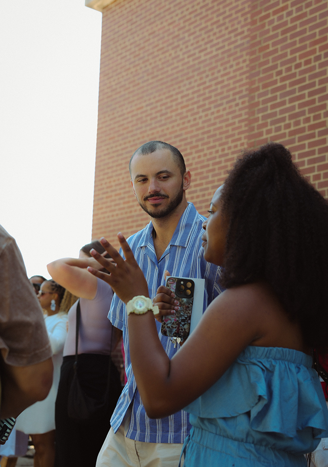 People chatting outdoors by a brick wall, one person holding a phone and another in a blue ruffled top.