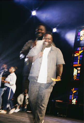 Two men on a stage under bright lights, one blurred in motion in the foreground, stained-glass panels behind.