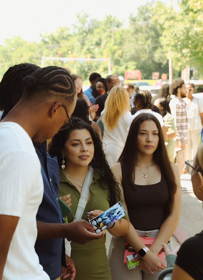 Three students talking outdoors at a crowded event, with one holding a colorful snack pack.