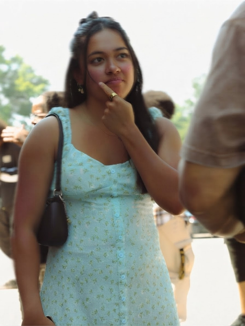 Woman in a light blue dress standing outdoors with hand on chin, looking thoughtful.