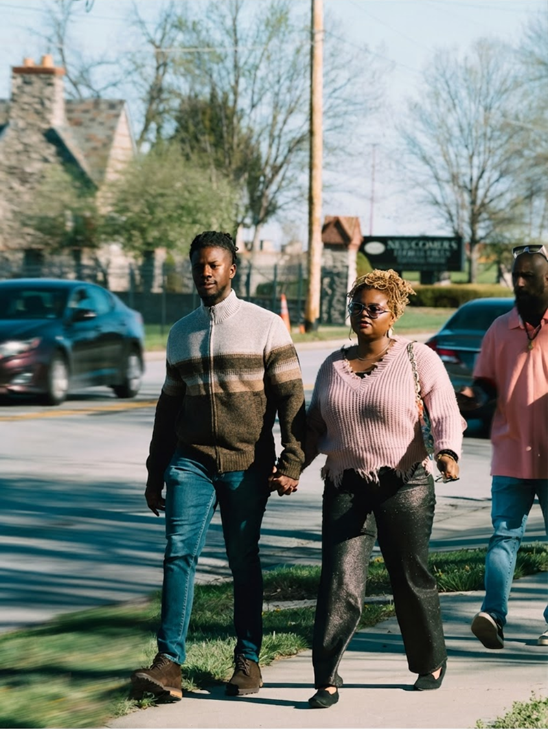 Three people walking on a suburban street, with trees, houses, and passing cars in the background.