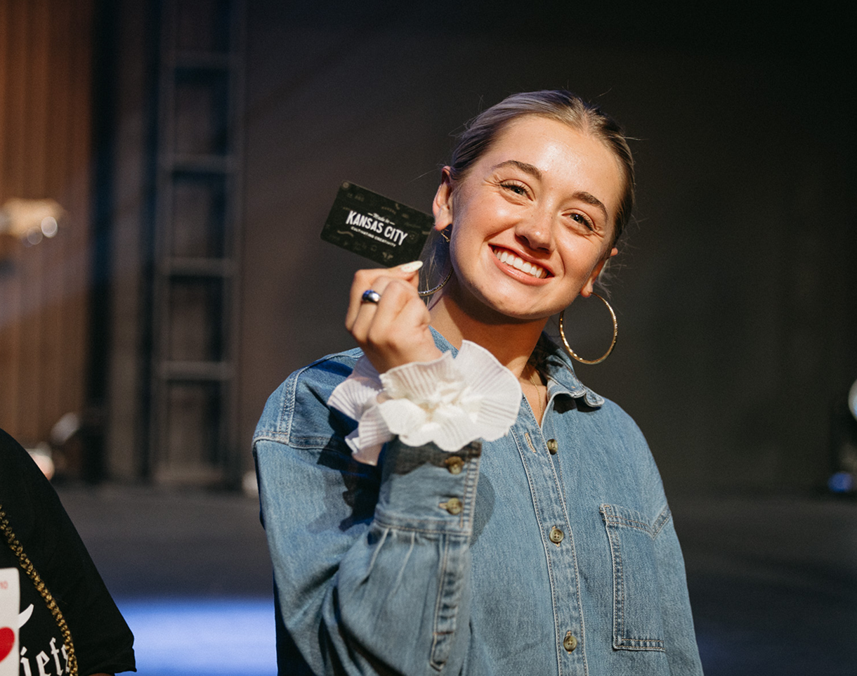 Smiling woman in denim jacket holding up a small card on a dimly lit stage