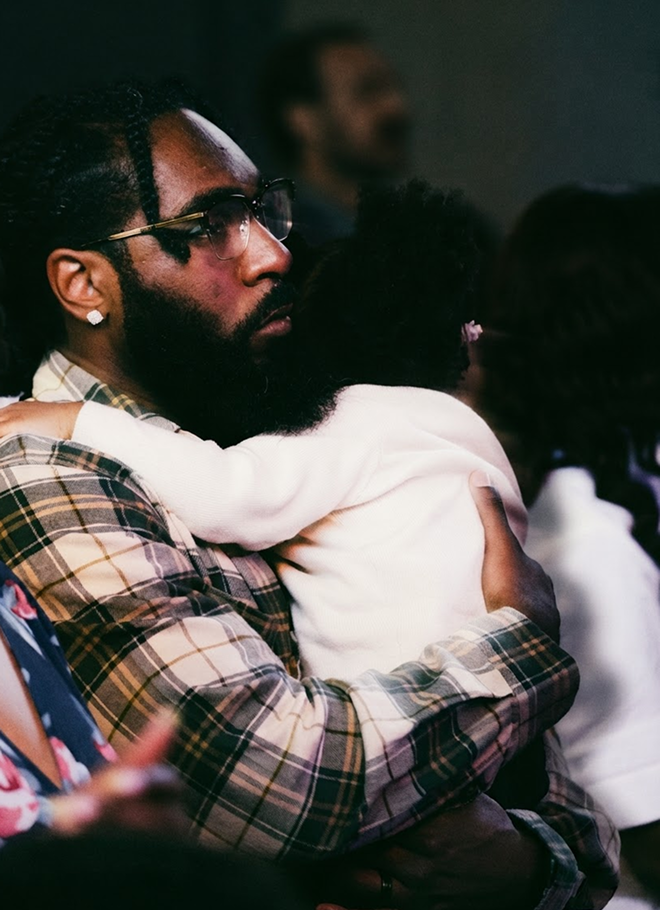 Man in plaid shirt and glasses holding a child in a white outfit indoors