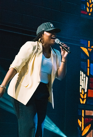Singer performing on stage with microphone, wearing a white blazer and green cap under blue lighting.