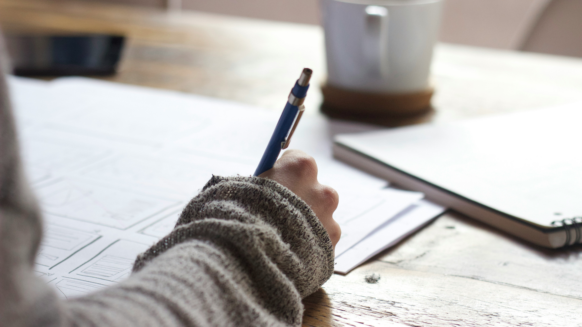 Person writing with a pen at a desk beside papers and a laptop