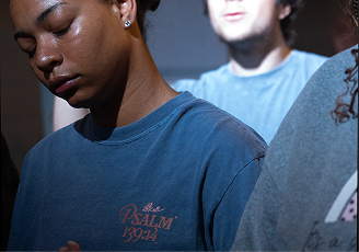 Three people in blue shirts sit in dim light, one in the foreground with eyes closed.