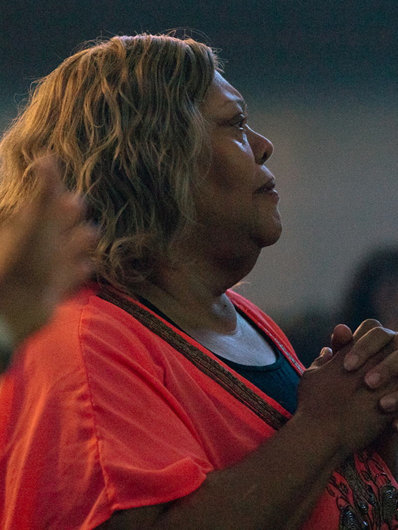 Person in an orange shirt standing with hands clasped, appearing to pray or sing in a dim indoor setting