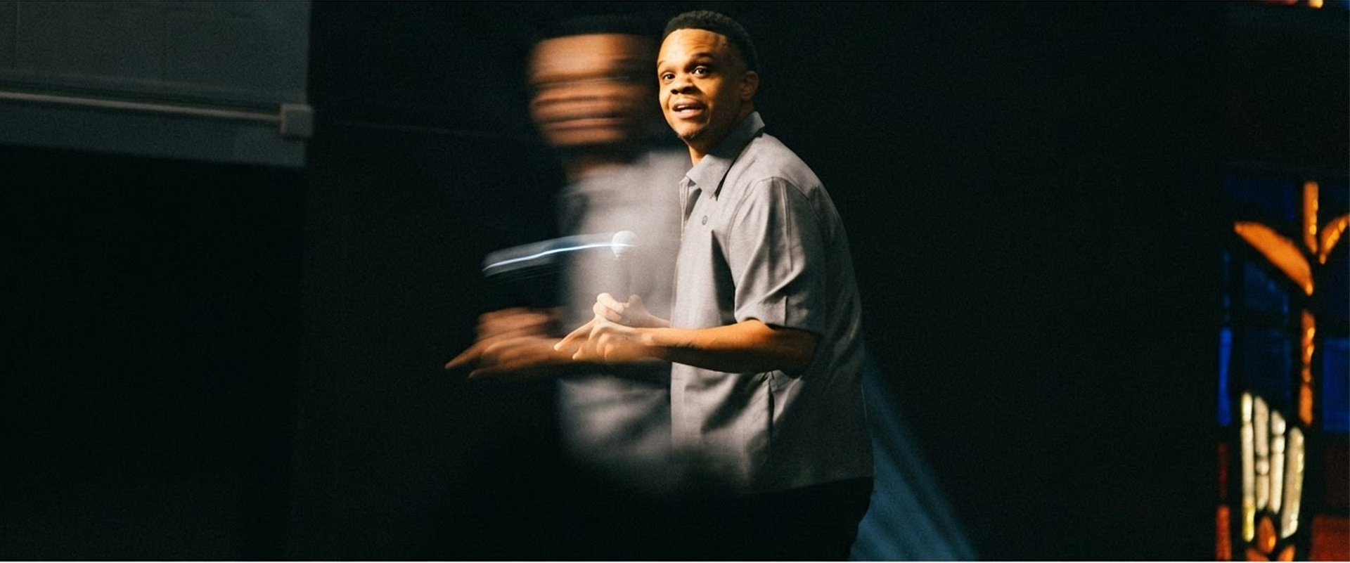 Speaker on a dark stage, gesturing mid-speech with warm spotlight and blurred background