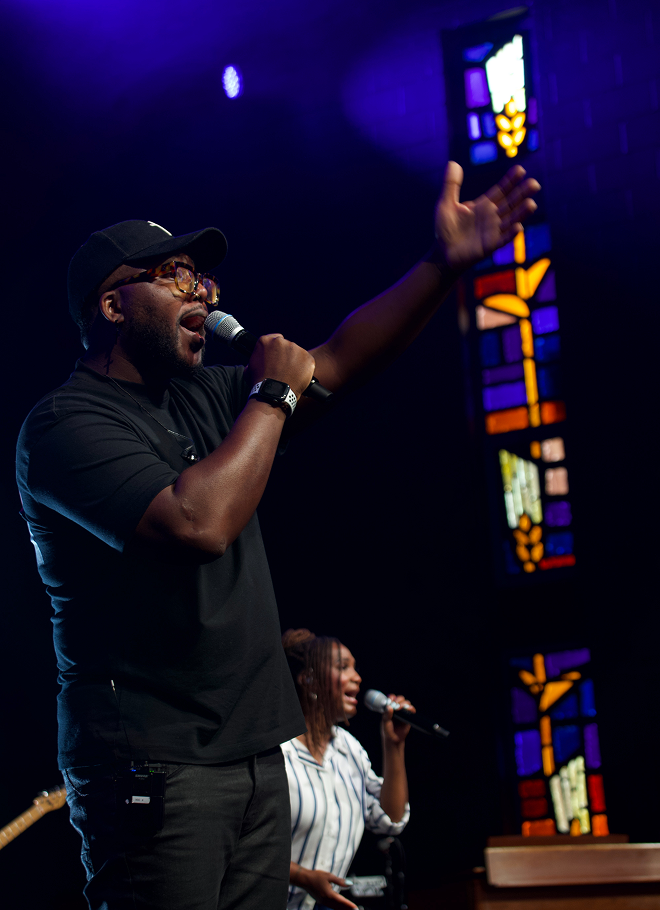 Singer on stage in black cap and shirt, holding a mic and raising an arm under purple lights.