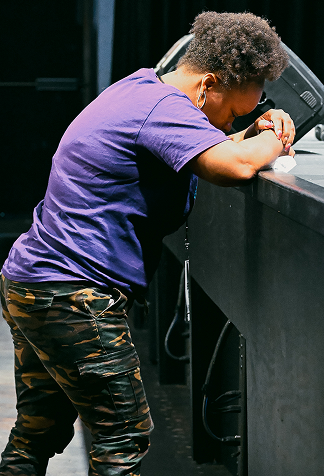 Person leaning on a black barrier, head resting on folded arms, wearing a purple shirt and camo pants.