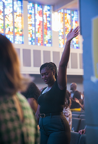 Woman raising one arm during a gathering in a church, with stained glass windows in the background.