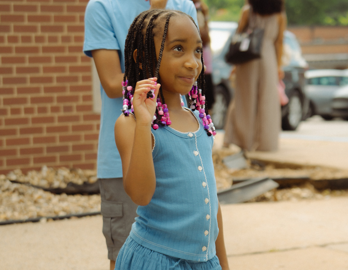 Girl in blue dress holding up two fingers outdoors near a brick building