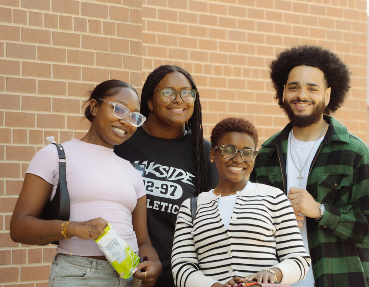 Four friends smiling in front of a brick wall, one holding a snack bag.