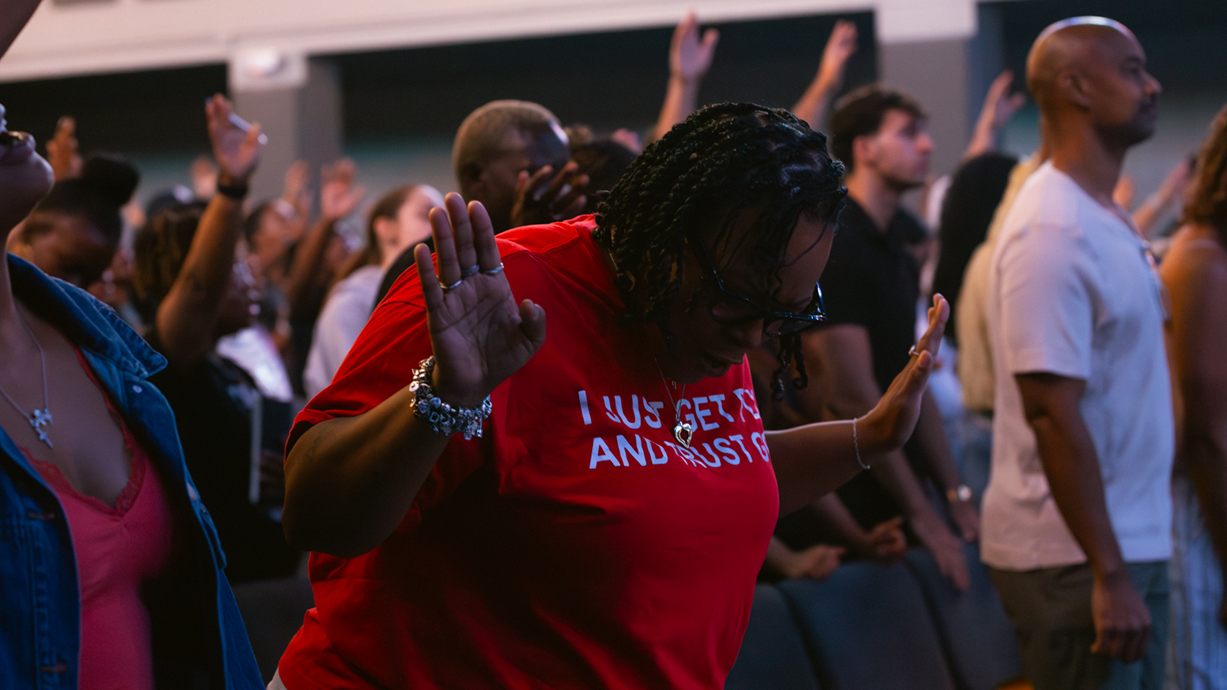 Crowd worshiping indoors, people raising hands in prayer and singing.