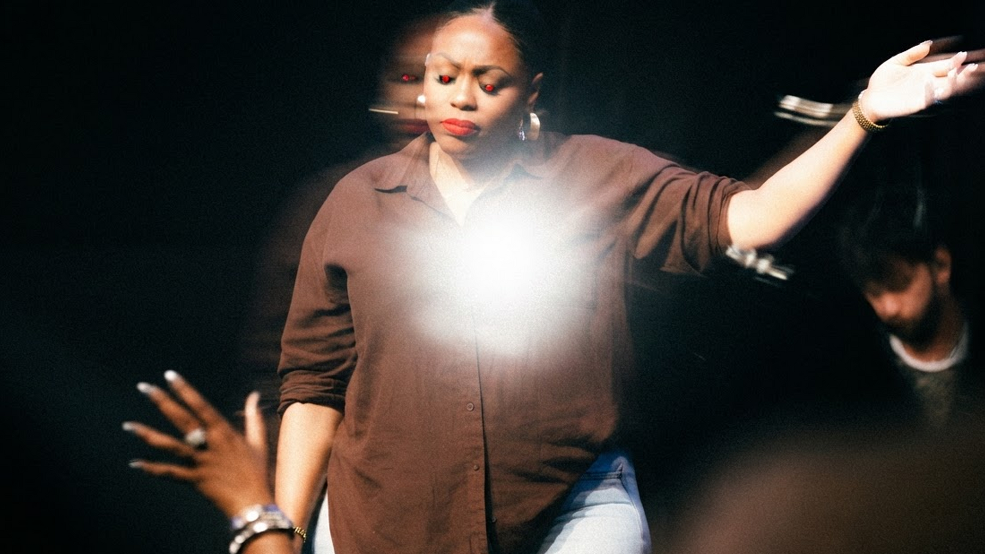 Singer performing on stage with arms raised, spotlight at center, audience hands reaching up in foreground