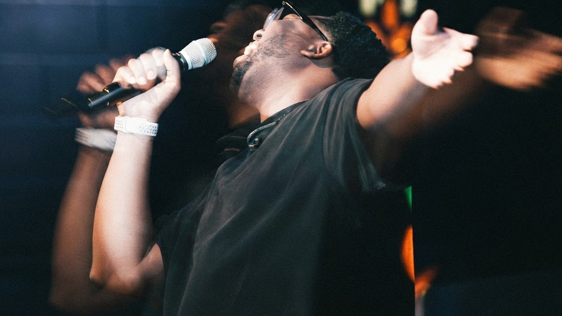 Singer performing on stage, holding a microphone with arms raised under warm concert lights