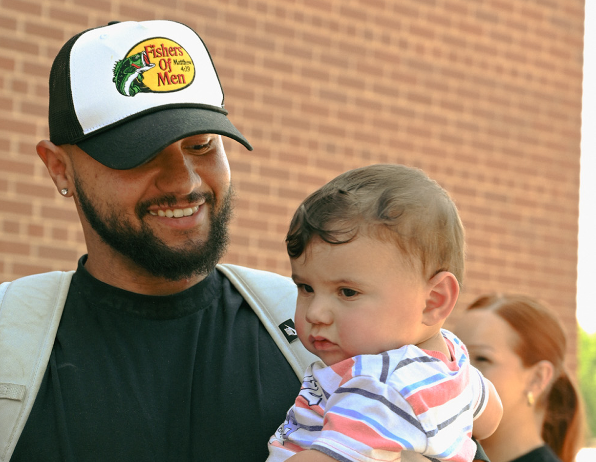 Man holding a baby outdoors, both wearing casual clothes, with a brick wall in the background