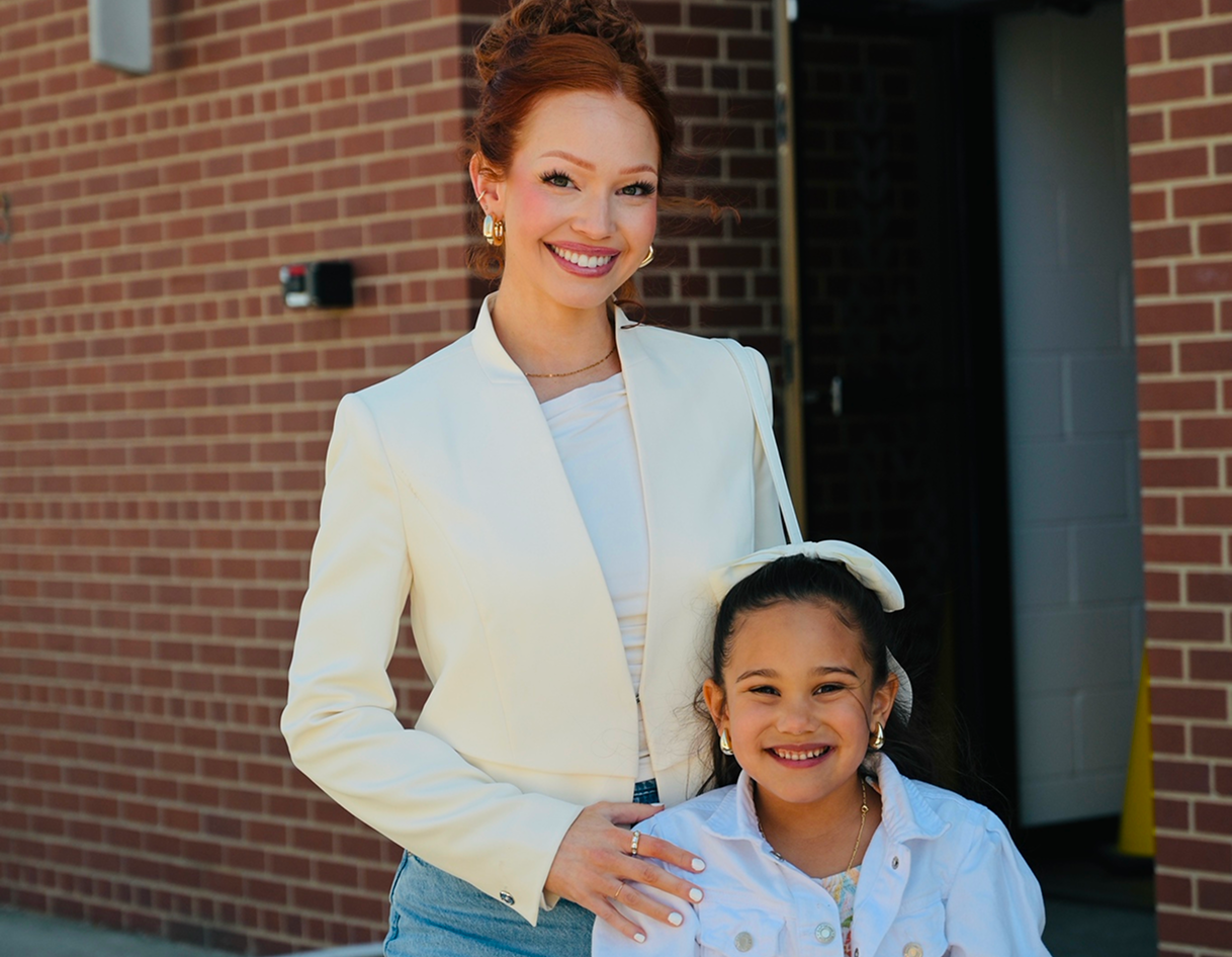 Two people smiling outside a brick building, one standing behind the other with a hand on her shoulder.