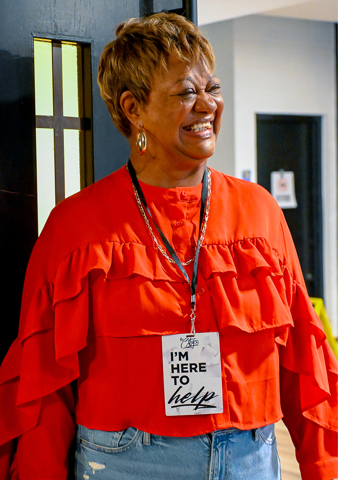 Smiling woman in a bright red blouse with a name tag, standing indoors near a doorway.