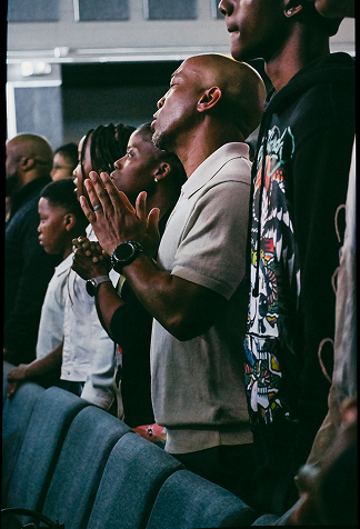 People standing and praying in a church, hands clasped near blue pews.