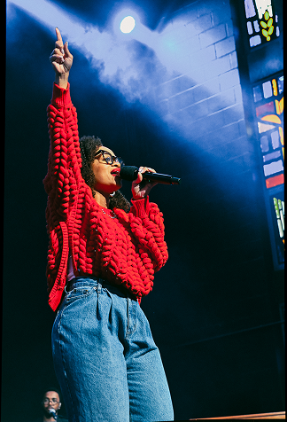 Singer in a red sweater and jeans performing onstage, holding a microphone and raising one hand.