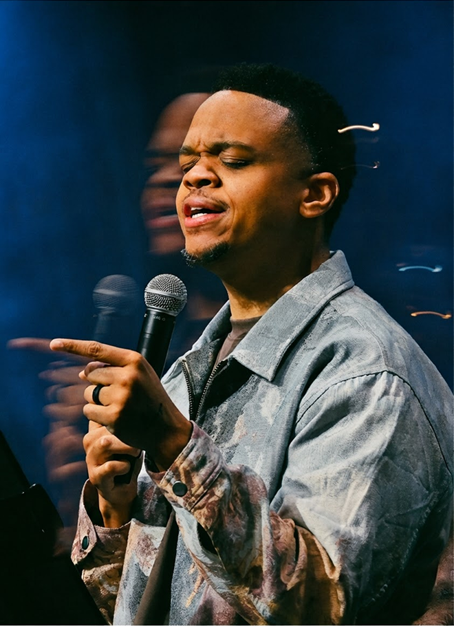 Person speaking into a microphone, pointing, on a stage with blue lighting and a blurred background