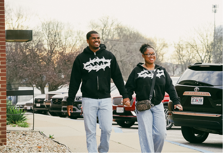Two people in matching black hoodies and jeans walking hand in hand on a sidewalk near parked cars