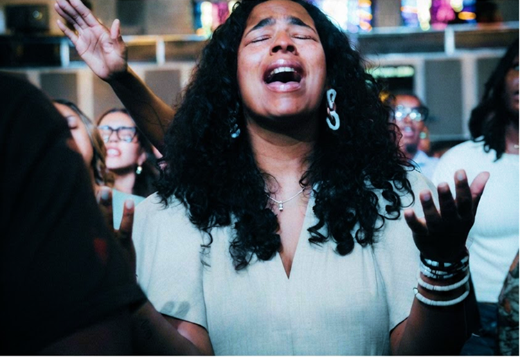 Woman with curly hair singing with eyes closed and hands raised in a lively indoor crowd scene