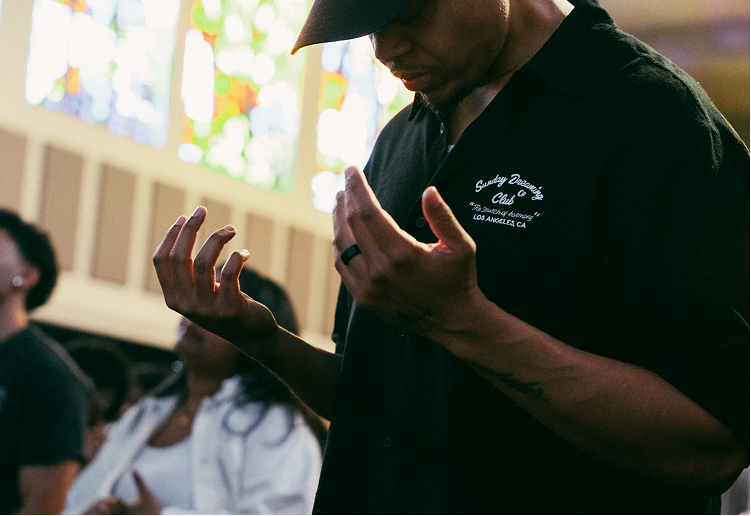 Person in black shirt with head bowed and hands raised in a church, stained glass visible in the background