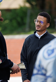 Man in glasses and black sweater speaking outdoors, with two people in the foreground.